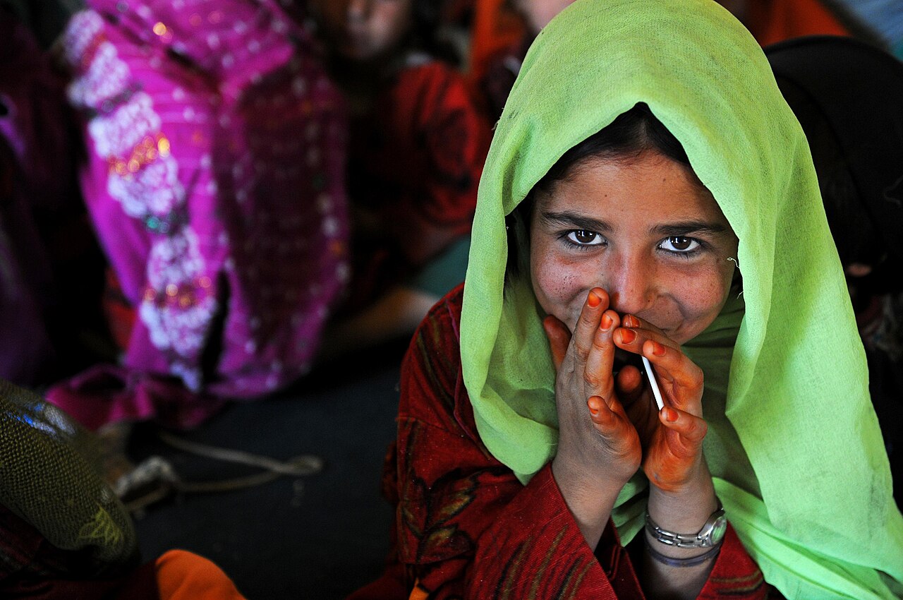 A young Afghan girl in a green scarf holds a pen, hands clasped over her mouth